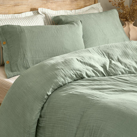 Lark Cotton Plain Duvet Cover Set in eucalyptus green, crinkled muslin cotton with wooden button fastening, styled on a bed with striped pillowcases in a bedroom setting.