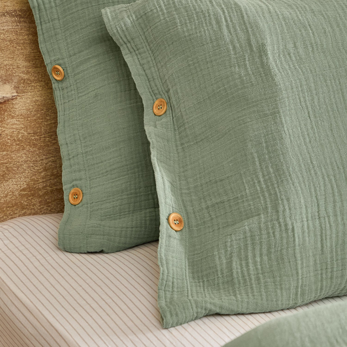Close-up of the Lark Cotton Plain Duvet Cover Set in eucalyptus green, showing crinkled muslin cotton pillow shams with wooden button fastenings on a striped bed sheet.