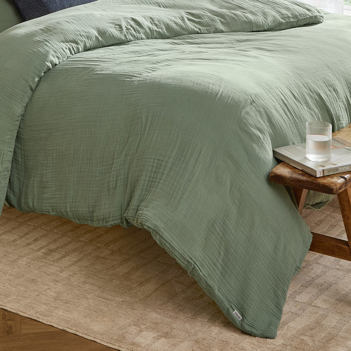 Lark Cotton Plain Duvet Cover Set in eucalyptus green, crinkled muslin cotton texture, shown on a bed beside a wooden stool with a glass and magazine.
