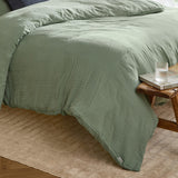 Lark Cotton Plain Duvet Cover Set in eucalyptus green, crinkled muslin cotton texture, shown on a bed beside a wooden stool with a glass and magazine.