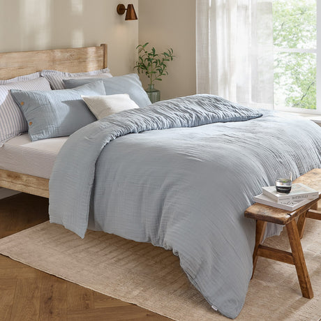 Lark Cotton Plain Duvet Cover Set in Mineral Blue with crinkled muslin texture, styled on a bed with striped pillowcases in a wood-framed bedroom setting.