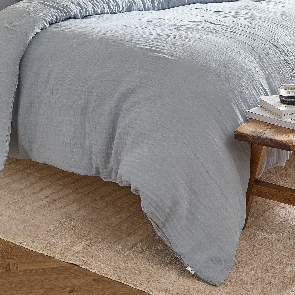 Lark Cotton Plain Duvet Cover Set in Mineral Blue on a bed, showing the crinkled double-layer muslin texture, styled with a wooden stool and woven rug in a bedroom setting.
