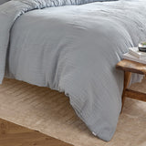 Lark Cotton Plain Duvet Cover Set in Mineral Blue on a bed, showing the crinkled double-layer muslin texture, styled with a wooden stool and woven rug in a bedroom setting.