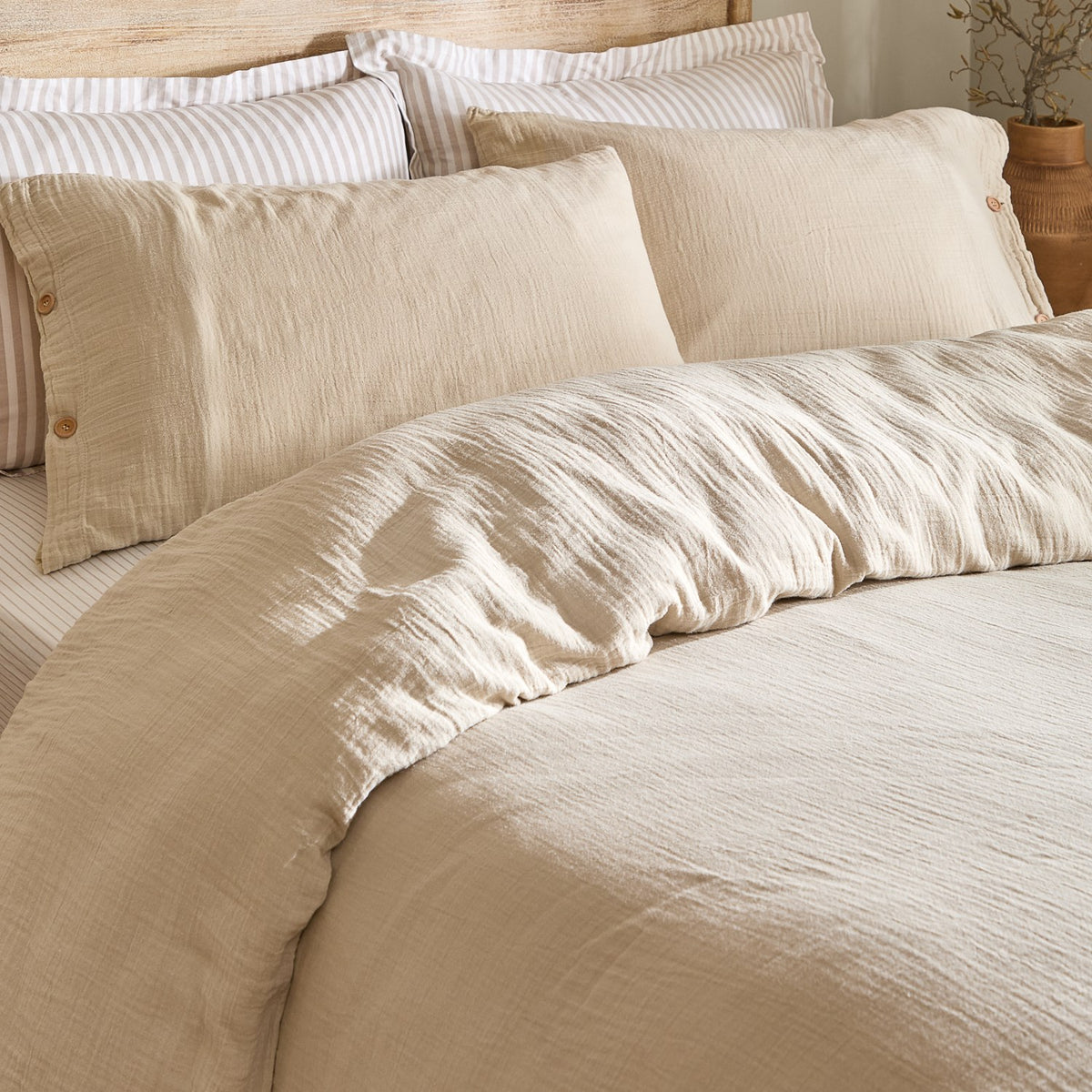 Lark Cotton Plain Duvet Cover Set in Natural, close-up on a bed showing crinkled muslin cotton texture with matching buttoned pillowcases and striped pillows behind.