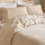 Lark Cotton Plain Duvet Cover Set in Natural, close-up on a bed showing crinkled muslin cotton texture with matching buttoned pillowcases and striped pillows behind.