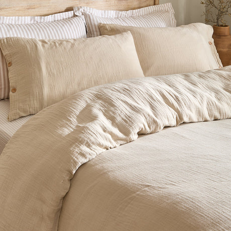 Lark Cotton Plain Duvet Cover Set in Natural, close-up on a bed showing crinkled muslin cotton texture with matching buttoned pillowcases and striped pillows behind.