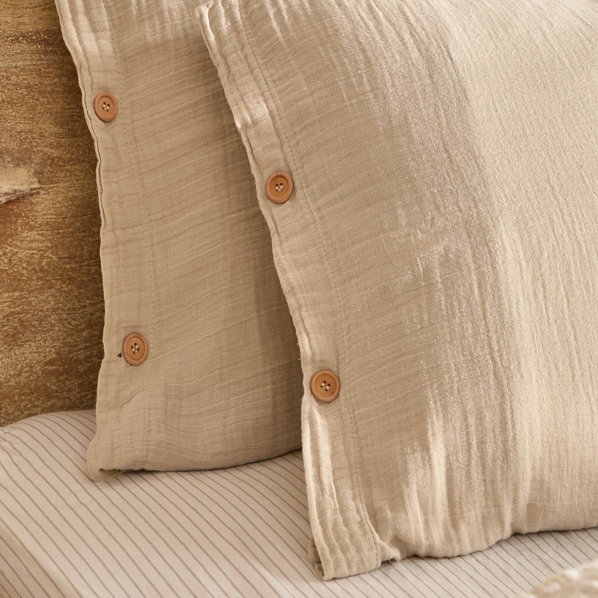 Lark Cotton Plain Duvet Cover Set in natural, close-up of crinkled muslin cotton pillowcases with button fastenings, styled on a striped bed sheet.