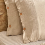 Lark Cotton Plain Duvet Cover Set in natural, close-up of crinkled muslin cotton pillowcases with button fastenings, styled on a striped bed sheet.