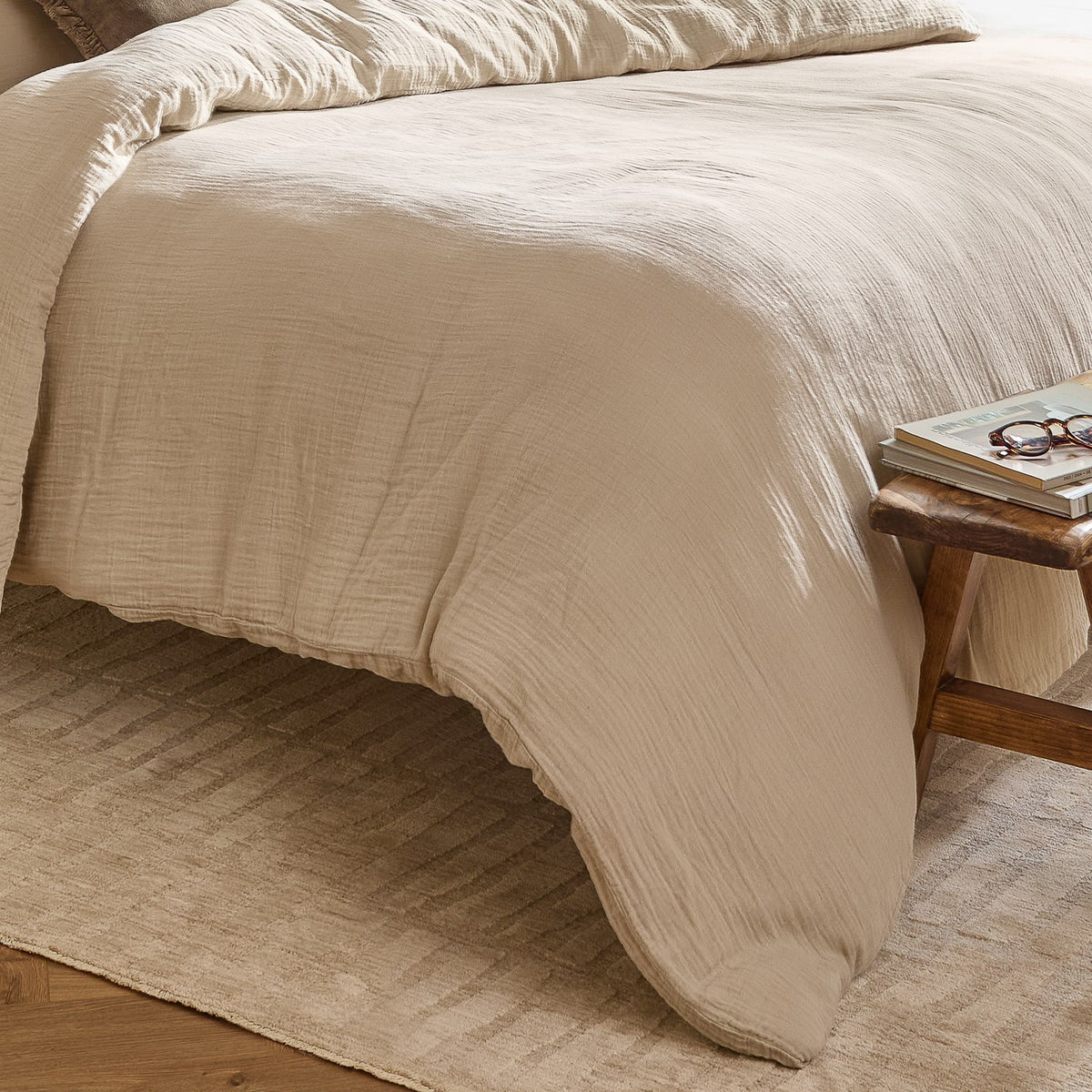 Lark Cotton Plain Duvet Cover Set in natural beige, crinkled muslin cotton texture, styled on a bed beside a wooden stool with glasses and magazines.