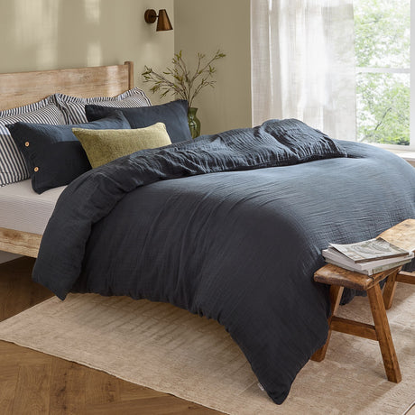 Lark Cotton Plain Duvet Cover Set in navy on a wooden bed, showing crinkled cotton texture with striped and olive accent pillows in a bedroom setting.