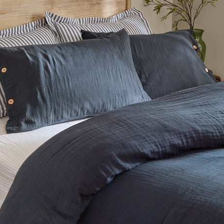 Lark Cotton Plain Duvet Cover Set in navy shown on a bed with matching pillowcases and striped bedding, crinkled cotton muslin texture and button fastenings visible.