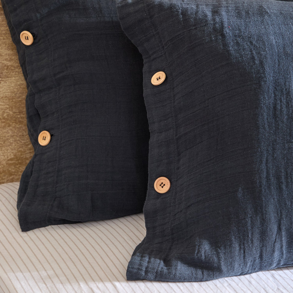 Lark Cotton Plain Duvet Cover Set in navy, close-up of crinkled muslin-weave fabric with wooden buttons on pillowcase, styled on a bed with striped sheeting.