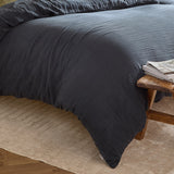 Lark Cotton Plain Duvet Cover Set in navy, shown on a bed with a crinkled cotton muslin texture, styled beside a wooden stool and stacked books on a woven rug.