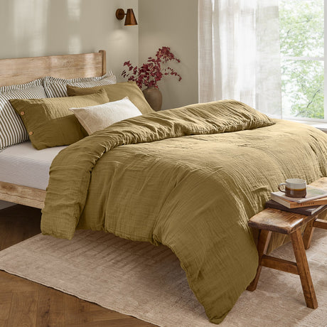 Lark Cotton Plain Duvet Cover Set in olive on a bed with crinkled muslin texture and wooden button-fastened pillowcases, shown in a bedroom with striped accent pillows.