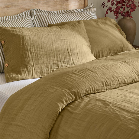 Lark Cotton Plain Duvet Cover Set in Olive, showing crinkled muslin cotton duvet and pillowcases with wooden button fastenings, styled on a bed with striped pillows.