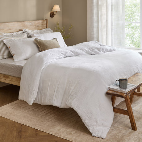 Lark Cotton Plain Duvet Cover Set in white with crinkled muslin texture on a wooden bed, styled with striped and velvet cushions in a neutral bedroom.