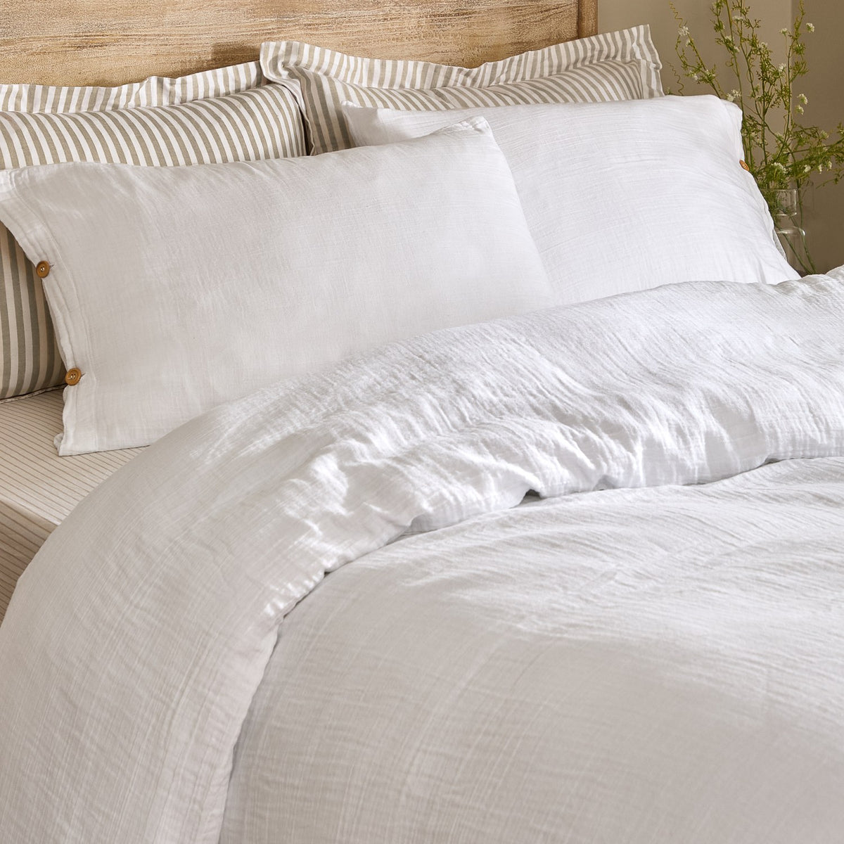 Lark Cotton Plain Duvet Cover Set in white on a bed with matching pillowcases and striped scatter cushions against a wooden headboard, showing the crinkled muslin texture.