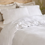 Lark Cotton Plain Duvet Cover Set in white on a bed with matching pillowcases and striped scatter cushions against a wooden headboard, showing the crinkled muslin texture.