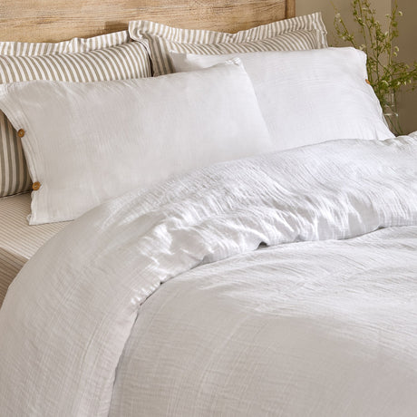 Lark Cotton Plain Duvet Cover Set in white on a bed with matching pillowcases and striped scatter cushions against a wooden headboard, showing the crinkled muslin texture.