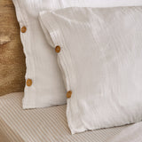 Close-up of the Lark Cotton Plain Duvet Cover Set in white, showing crinkled muslin cotton texture and buttoned pillowcase openings with wooden buttons on a striped bed base.