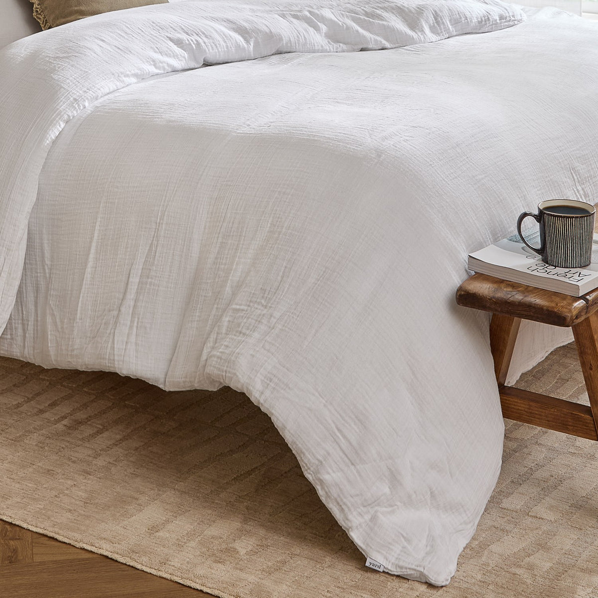 Lark Cotton Plain Duvet Cover Set in white on a bed with crinkled muslin texture, styled in a bedroom with a wooden stool, mug and magazine alongside.