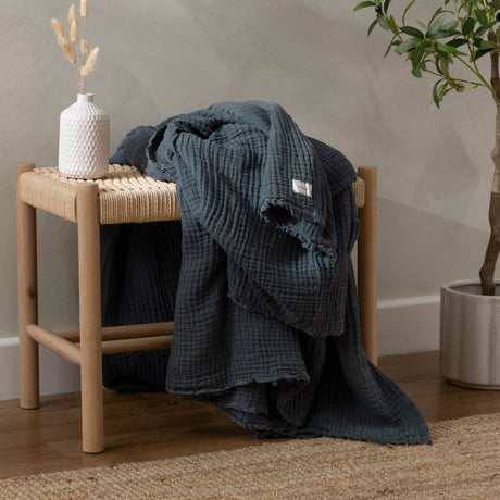 Lark Plain Cotton Throw in Dusk blue, crinkled cotton muslin with frayed edges, draped over a wooden bench beside a vase and plant.