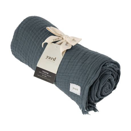 Lark Plain Cotton Throw in dusk blue, rolled up with frayed edge visible, textured cotton muslin fabric and cream branded tag tied with ribbon, shown on a plain white background.