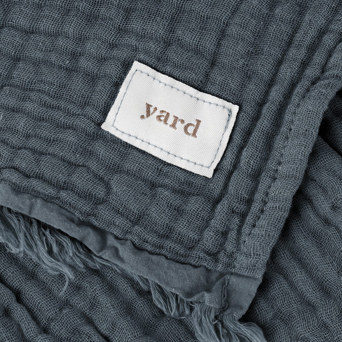 Close-up of the Lark Plain Cotton Throw in dusk blue, showing crinkled cotton muslin texture, frayed edge, and a white "yard" fabric label.