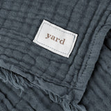 Close-up of the Lark Plain Cotton Throw in dusk blue, showing crinkled cotton muslin texture, frayed edge, and a white "yard" fabric label.