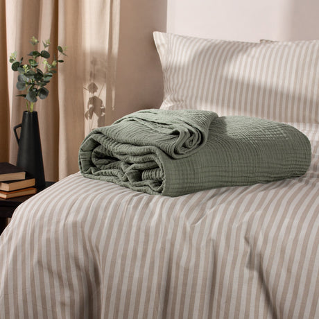 Lark Plain Cotton Throw in eucalyptus green, folded with crinkled cotton muslin texture on a beige striped bed with matching pillow.