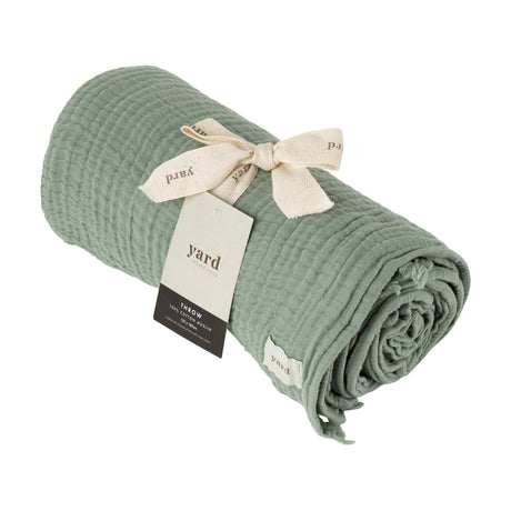 Lark Plain Cotton Throw in eucalyptus green, rolled with a crinkled muslin texture, tied with a cream ribbon and branded Yard tag, on a white background.