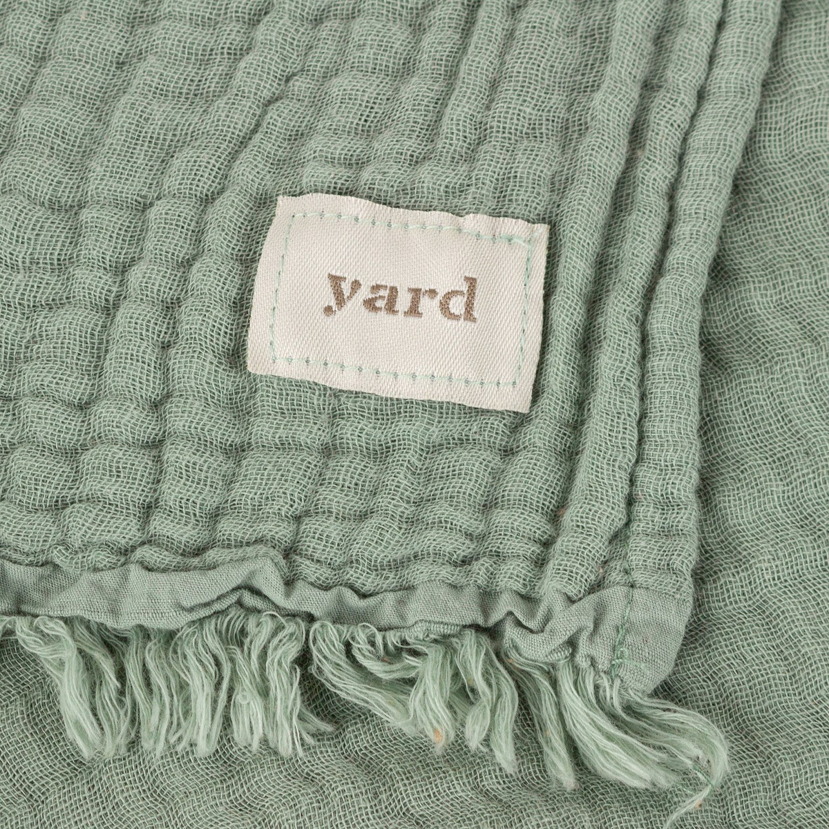 Close-up of the Lark Plain Cotton Throw in eucalyptus green, showing crinkled muslin texture, woven fabric label, and knotted fringe edge.