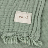 Close-up of the Lark Plain Cotton Throw in eucalyptus green, showing crinkled muslin texture, woven fabric label, and knotted fringe edge.
