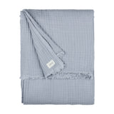 Lark Plain Cotton Throw in mineral blue, folded showing crinkled muslin texture and frayed edge with fabric label, on a plain white background.