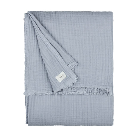 Lark Plain Cotton Throw in mineral blue, folded showing crinkled muslin texture and frayed edge with fabric label, on a plain white background.
