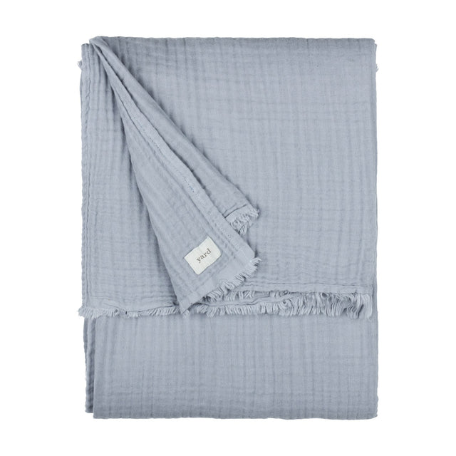Lark Plain Cotton Throw in mineral blue, folded showing crinkled muslin texture and frayed edge with fabric label, on a plain white background.