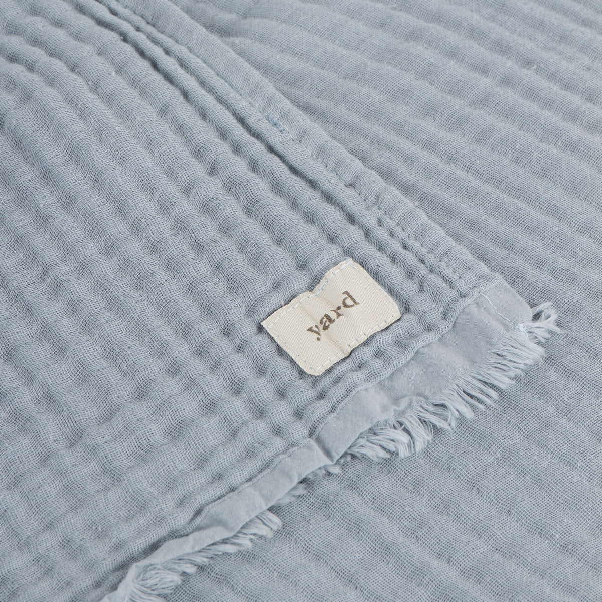 Close-up of the Lark Plain Cotton Throw in mineral blue, showing crinkled muslin texture, frayed edge trim, and a woven "yard" fabric label.