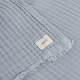 Close-up of the Lark Plain Cotton Throw in mineral blue, showing crinkled muslin texture, frayed edge trim, and a woven "yard" fabric label.