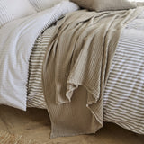 Lark Plain Cotton Throw in mole draped over a striped duvet bed, showing its textured cotton muslin weave and frayed edge finish.