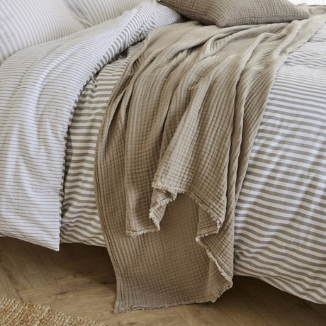 Lark Plain Cotton Throw in mole draped over a striped duvet bed, showing its textured cotton muslin weave and frayed edge finish.