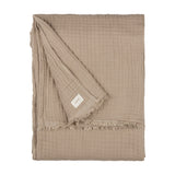 Lark Plain Cotton Throw in mole, a folded four-layer cotton muslin throw with a fringed edge and fabric brand tag, shown on a plain white background.