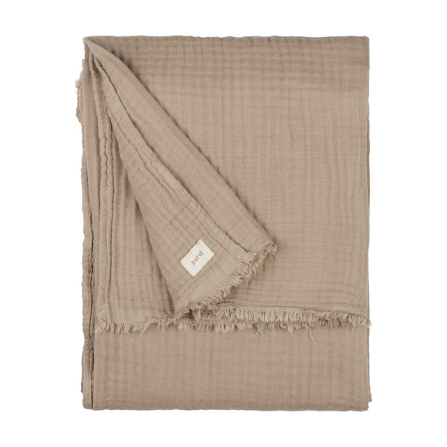 Lark Plain Cotton Throw in mole, a folded four-layer cotton muslin throw with a fringed edge and fabric brand tag, shown on a plain white background.