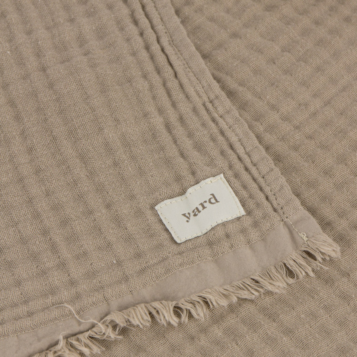 Close-up of the Lark Plain Cotton Throw in mole, showing a waffle-textured cotton weave with stitched hem, woven "yard" label, and fringed edge.
