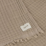 Close-up of the Lark Plain Cotton Throw in mole, showing a waffle-textured cotton weave with stitched hem, woven "yard" label, and fringed edge.