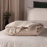 Lark Plain Cotton Throw in natural beige, folded on a bed showing crinkled muslin texture and frayed edge, styled with neutral bedding and cushions.