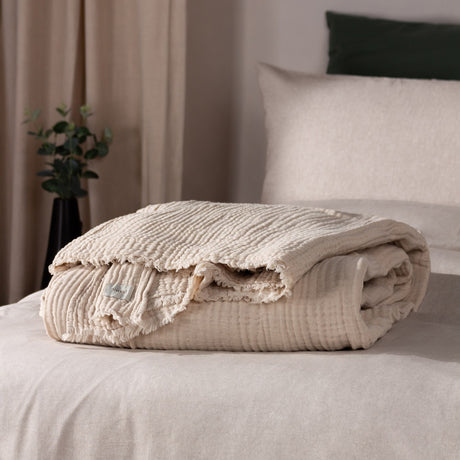 Lark Plain Cotton Throw in natural beige, folded on a bed showing crinkled muslin texture and frayed edge, styled with neutral bedding and cushions.