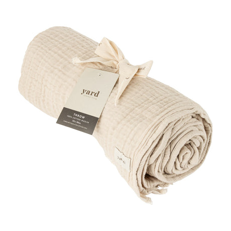 Lark Plain Cotton Throw in Natural, shown rolled with a crinkled muslin texture and Yard branded tag, against a plain white background.