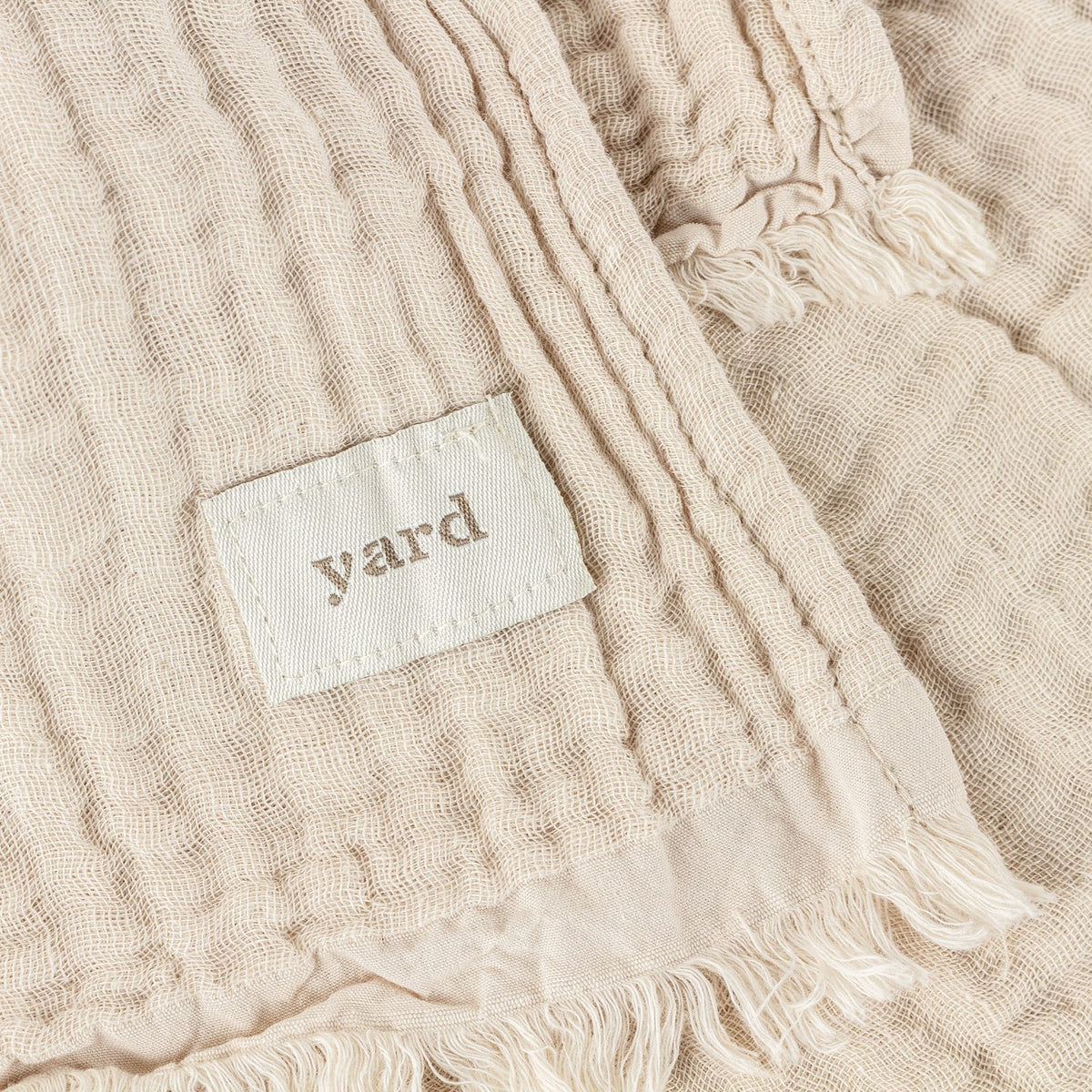 Close-up of the Lark Plain Cotton Throw in natural beige, showing crinkled muslin texture, frayed edge fringing, and a woven "yard" label.