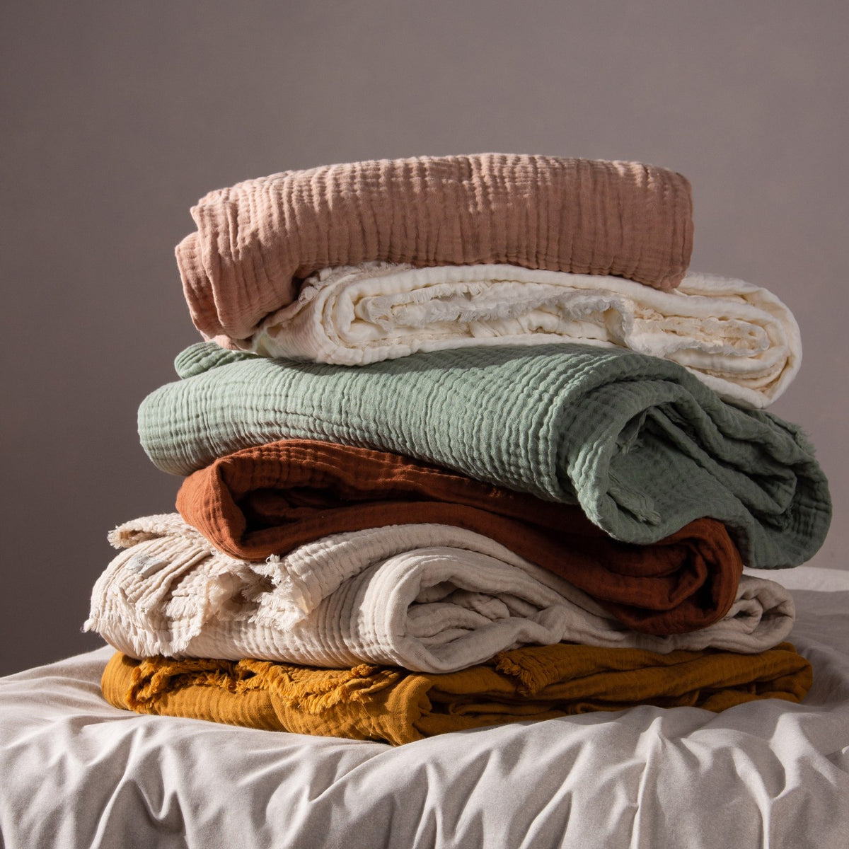 Stack of folded cotton muslin throws in blush pink, cream, sage green, rust, beige, and mustard, including the Lark Plain Cotton Throw in Natural, on a bed against a grey background.