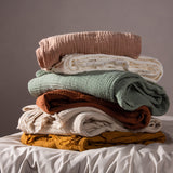 Stack of folded cotton muslin throws in blush pink, cream, sage green, rust, beige, and mustard, including the Lark Plain Cotton Throw in Natural, on a bed against a grey background.